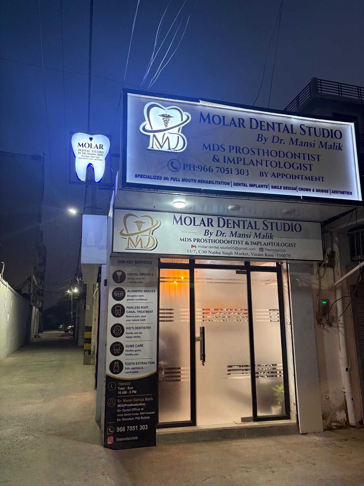 Molar Dental Studio after dark: both lit signboards, the tooth-shaped sign out over the lane, and the key-services panel beside the glass door